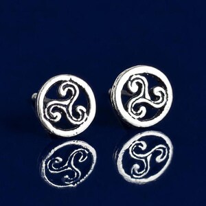 May include: A pair of silver-toned stud earrings featuring a circular frame with an openwork triskelion design. The earrings have a polished metallic finish and are displayed against a dark blue background with a clear reflection visible below.
