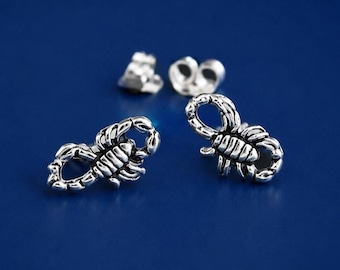 May include: A pair of silver-toned stud earrings shaped like scorpions with oxidised detailing to highlight the segmented body and curved tail. The earrings include two matching metal butterfly-style earring backs positioned behind the studs.
