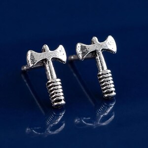 May include: A pair of silver-colored axe-shaped stud earrings. Each earring features a detailed axe head and a textured handle. The earrings are displayed on a reflective blue surface.