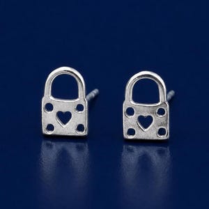 May include: A pair of silver-toned stud earrings shaped like padlocks. Each earring features a cutout heart in the center and four small circular perforations at the corners of the square base, with a rounded arch top and a post backing.