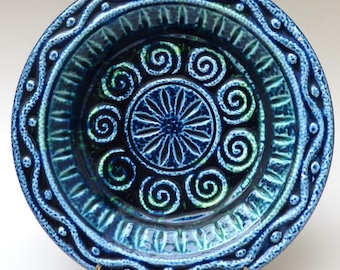 May include: A blue ceramic plate with a raised design of spirals and a central flower. The plate has a textured rim with a wavy pattern.