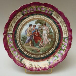 May include: A decorative porcelain plate with a scalloped edge and a pink and gold rim. The center of the plate features a hand-painted scene of four figures in a garden setting.