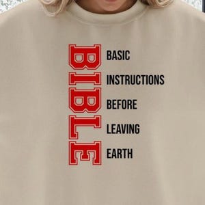 Puede incluir: Sudadera beige con la palabra "BIBLE" en letras rojas apiladas verticalmente a la izquierda. Las palabras "BASIC INSTRUCTIONS BEFORE LEAVING EARTH" están impresas en negro a la derecha de "BIBLE".