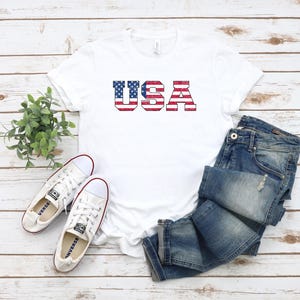 以下が含まれることがあります： 赤、白、青で「USA」とプリントされた白いTシャツ、白いスニーカー、青いジーンズ。 Tシャツのデザインはアメリカ国旗に似ています。明るい木目調の表面に配置されています。