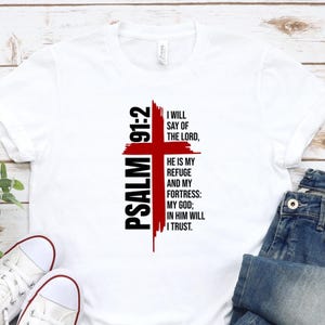 May include: A white short-sleeved t-shirt featuring a red cross graphic with black text that reads: PSALM 91:2 I WILL SAY OF THE LORD, HE IS MY REFUGE AND MY FORTRESS: MY GOD; IN HIM WILL I TRUST. The design is centered on the front of the shirt.