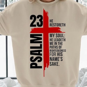 May include: A beige sweatshirt with a red cross design. The text "23 PSALM" is on the left side. The cross is overlaid with the words "HE RESTORETH" and a religious quote in black. The design is in red and black.