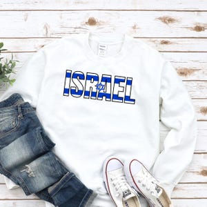 Könnte beinhalten: Ein weißes langärmeliges Sweatshirt mit Rundhalsausschnitt und dem Wort ISRAEL in fetter Blockschrift. Die Buchstaben sind mit horizontalen blauen und weißen Streifen gefüllt, und ein blauer Davidstern ersetzt den Buchstaben A.