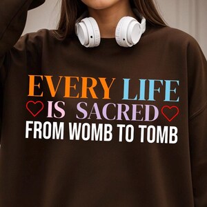 Puede incluir: Una sudadera marrón con el texto "EVERY LIFE IS SACRED FROM WOMB TO TOMB" en letras coloridas. Las palabras son de color naranja, azul, morado y blanco. Unos auriculares blancos descansan sobre los hombros.