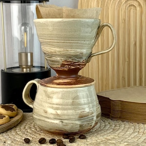 May include: A ceramic coffee pour-over set with a brown base and a light beige top, on a matching mug. Coffee filters are in the pour-over. A black lantern with a gold handle and a wooden serving tray with biscuits are also in the image.