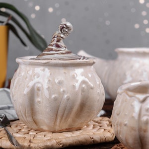 May include: A white ceramic sugar bowl with a decorative lid and a unique swirl design. The bowl has a textured surface and is sitting on a woven placemat.