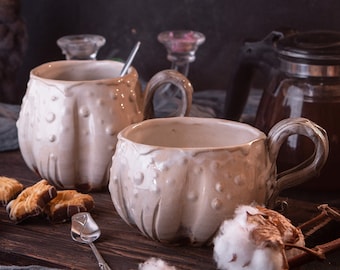 May include: Two white ceramic mugs with textured surfaces and handles, filled with a brown liquid, on a wooden table with a blue cloth, a glass teapot, cotton bolls, and cookies.