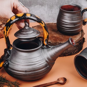 Può includere: Una teiera nera e gialla con un coperchio e una tazza abbinata. La teiera e la tazza sono in ceramica e hanno una finitura testurizzata.