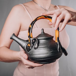 Puede incluir: Una tetera de cerámica negra con un asa única con un patrón amarillo y negro. La tetera tiene un cuerpo redondeado y una tapa con un pequeño pomo.