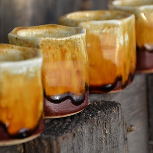 Peut inclure: Rangée de tasses en céramique faites à la main avec un glaçage unique. Les tasses ont une forme carrée aux bords arrondis. Le glaçage passe du blanc crème en haut à des nuances de jaune et de brun, se terminant par une base marron foncé.