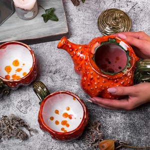 May include: Ceramic teapot and mugs set. The teapot is orange with a textured surface and a green handle. The mugs are red with green handles and white interiors with orange spots. The set is designed for serving tea or other beverages.