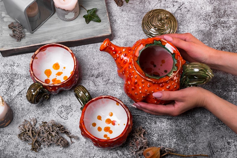 May include: Ceramic teapot and mugs set. The teapot is orange with a textured surface and a green handle. The mugs are red with green handles and white interiors with orange spots. The set is designed for serving tea or other beverages.