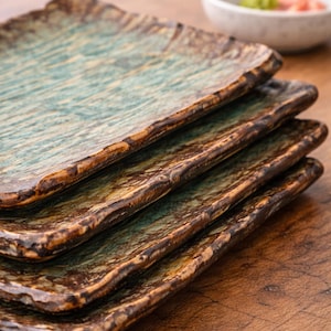 Könnte beinhalten: Ein Stapel von vier rechteckigen Keramikplatten mit einem rustikalen, strukturierten Design. Die Platten haben eine türkis-braune Farbgebung mit einer rauen, unebenen Kante. Im Hintergrund befindet sich eine kleine Schüssel mit grünem und rosa Inhalt.