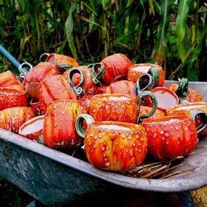 May include: A wheelbarrow filled with ceramic mugs. The mugs are pumpkin-shaped, with orange and yellow coloring and white speckles. Each mug has a green handle. The wheelbarrow is gray and sits in a grassy area.