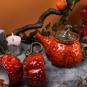 May include: A ceramic tea set with a teapot and two cups, all in a vibrant orange color with white spots. The teapot has a green handle and lid, and the cups have green handles. The set is displayed with candles and a decorative branch.