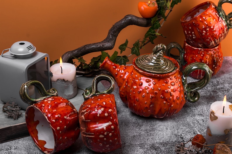 May include: A ceramic tea set with a teapot and two cups, all in a vibrant orange color with white spots. The teapot has a green handle and lid, and the cups have green handles. The set is displayed with candles and a decorative branch.