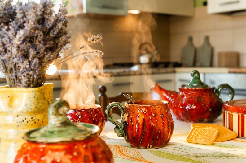 May include: A warm, inviting kitchen scene featuring a red ceramic teapot and mugs, with steam rising from the hot beverages. A yellow vase holds lavender, and cookies sit nearby. The scene evokes a cozy, relaxing atmosphere.