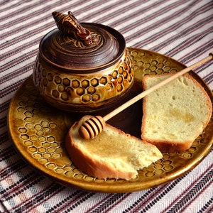 May include: A brown ceramic honey pot with a bee design on the lid, a wooden honey dipper, and two slices of bread with honey on a brown plate with a honeycomb design.