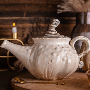 Può includere: Una teiera in ceramica bianca con una superficie testurizzata e un beccuccio unico. La teiera ha un coperchio con un pomo decorativo. La teiera è appoggiata su una superficie di legno.