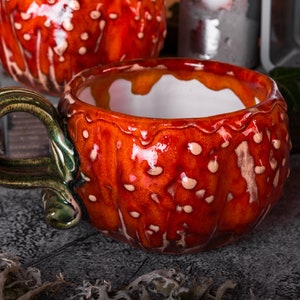 May include: A ceramic mug with a vibrant red and white spotted exterior, resembling a mushroom. The handle is a contrasting green and brown. The interior is white. The mug is designed with a unique, textured surface, perfect for a cozy beverage.
