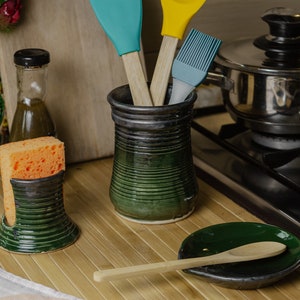 Pode incluir: Uma cena de cozinha com porta-utensílios de cerâmica, um porta-esponja e um descanso de colher. Os suportes são verde escuro com um design texturizado. Espátulas e um pincel em azul-petróleo e amarelo estão no porta-utensílios. Uma panela de aço inoxidável está no fogão.