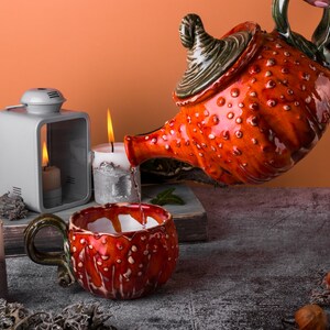 May include: A ceramic teapot and mug set with a vibrant red and white spotted design. The teapot is pouring liquid into the matching mug. Lit candles and a lantern create a warm, inviting atmosphere. The set is on a gray surface.