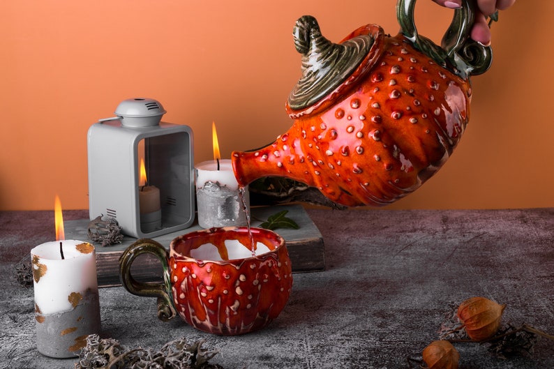 May include: A ceramic teapot and mug set with a vibrant red and white spotted design. The teapot is pouring liquid into the matching mug. Lit candles and a lantern create a warm, inviting atmosphere. The set is on a gray surface.