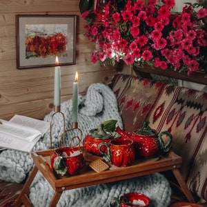 May include: A cozy scene featuring a wooden tray with a red ceramic tea set, including a teapot and cups, set on a chunky knit blanket. Two lit candles and a book are nearby. The background includes a floral arrangement and framed artwork.
