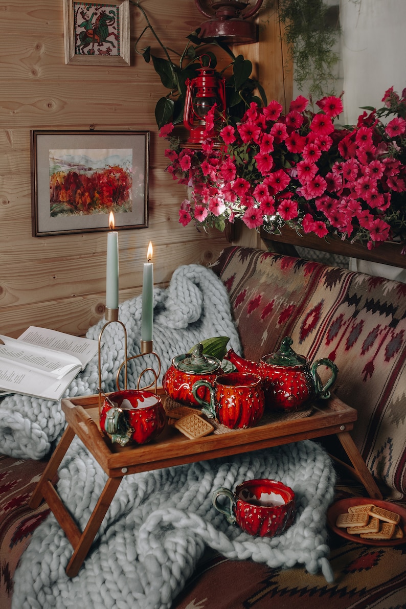 May include: A cozy scene featuring a wooden tray with a red ceramic tea set, including a teapot and cups, set on a chunky knit blanket. Two lit candles and a book are nearby. The background includes a floral arrangement and framed artwork.