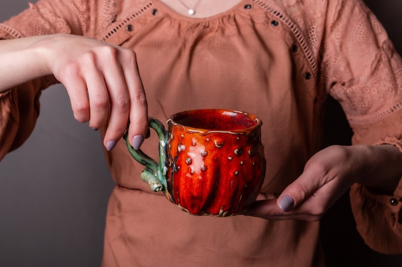 May include: A handmade ceramic mug with a vibrant red and orange exterior, accented with small, raised dots. The handle is a contrasting green, adding a unique touch to this handcrafted drinkware.