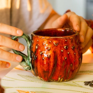 May include: A handmade ceramic mug with a textured, reddish-orange exterior and a green handle. The mug has a unique, organic shape with small, raised bumps. The interior is a darker shade of red. A spoon rests on the table next to the mug.