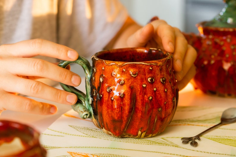 May include: A handmade ceramic mug with a textured, reddish-orange exterior and a green handle. The mug has a unique, organic shape with small, raised bumps. The interior is a darker shade of red. A spoon rests on the table next to the mug.