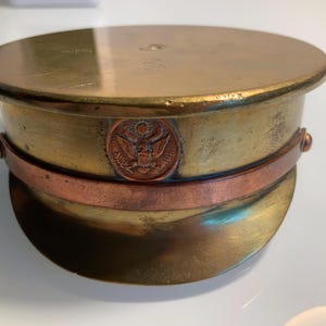Op de afbeelding: Een vintage militaire hoed van messing met een koperkleurige band en een cirkelvormig embleem met een adelaar. De hoed heeft een afgeronde bovenkant en een korte klep, met tekenen van slijtage. De kleur van de hoed is een mix van goud en brons.