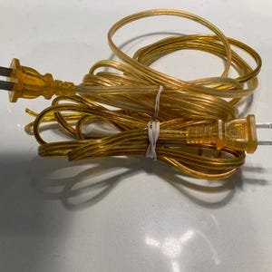 May include: A transparent yellow electrical cord with a two-prong plug. The cord is coiled and secured with a white twist tie. The plug and cord are made of a clear, amber-colored plastic.