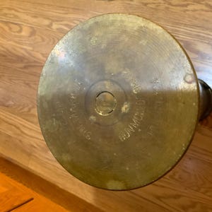 May include: A vintage brass artillery shell casing. The top of the shell is circular with engraved text including "MARK 14", "SIN-NIS", and "BUNMOU OF 124". The casing has a tarnished, aged appearance.