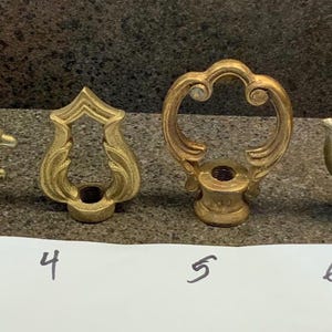 Könnte beinhalten: Eine Sammlung von sieben antiken Messing-Zierknöpfen, jeweils mit einem einzigartigen dekorativen Design. Die Zierknöpfe sind in einer Reihe angeordnet, mit einem weißen Papier darunter, das mit den Nummern 1 bis 7 beschriftet ist.