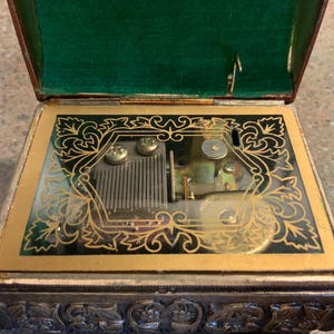 May include: An antique music box with a detailed silver-colored exterior and a hinged lid. The interior is lined with green velvet. The top features a glass panel with gold filigree and the mechanical components of the music box.