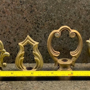 Pode incluir: Uma coleção de sete peças de ferragens decorativas antigas em latão, cada uma com um design ornamental único. As peças estão dispostas em fila, com uma fita métrica amarela por baixo. As ferragens são provavelmente para móveis ou armários.