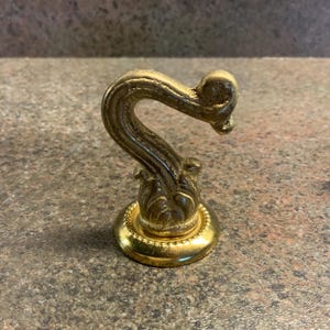 May include: A decorative, ornate brass hook with a curved design. The hook has a detailed floral base and a rounded top. The brass finish has a slightly aged appearance. A screw and a metal object are visible in the background.