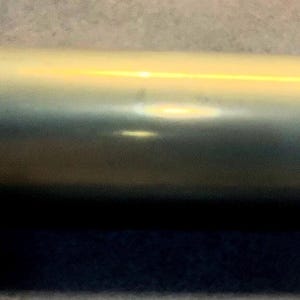 May include: A cylindrical brass shell casing with a smooth, polished metallic surface. The object features a slightly flared rim at one end and a flat, circular base at the other, maintaining a consistent tubular shape throughout its length.