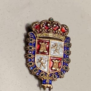 May include: A gold-toned, enameled brooch featuring a heraldic crest. The crest is divided into four sections with red, white, and blue enamel. The design includes a crown, towers, and figures. The brooch has a decorative border and a small animal at the bottom.