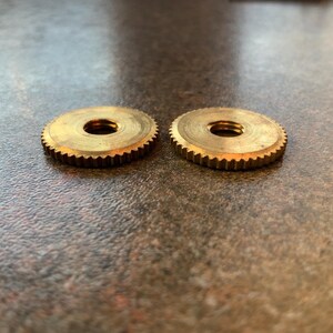 Pode incluir: Duas porcas rosqueadas de latão com um design circular e uma borda externa dentada. As porcas têm um orifício central e são posicionadas lado a lado em uma superfície texturizada. As porcas são douradas.