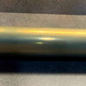 May include: A cylindrical brass object with a slightly tarnished finish. The object is a long tube with a flared end on one side and a flat end on the other. The brass has a golden hue with some darker areas.