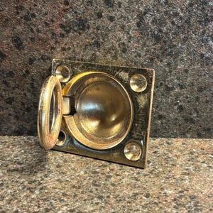 May include: A rectangular brass flush pull handle featuring a circular recessed center and a hinged ring pull. The metal surface displays visible scratches and a metallic gold finish, with four pre-drilled screw holes at each corner of the plate.