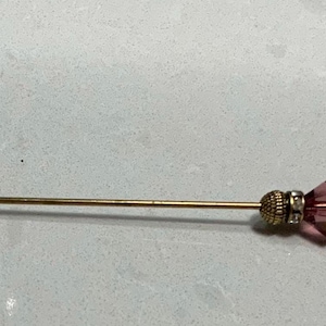 May include: A decorative hat pin with a gold-colored shaft and a dark purple, faceted glass head. The head is accented with clear crystals and a gold-colored metal band. The pin is designed for use with hats or other accessories.