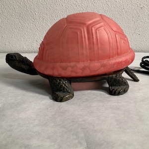 Op de afbeelding: Een vintage schildpadvormige lamp met een rode, getextureerde glazen schaal en een donkerbronzen voet. De lamp heeft een zwart netsnoer en is ontworpen om op een vlakke ondergrond te staan. De lamp is een decoratieve verlichtingsarmatuur.